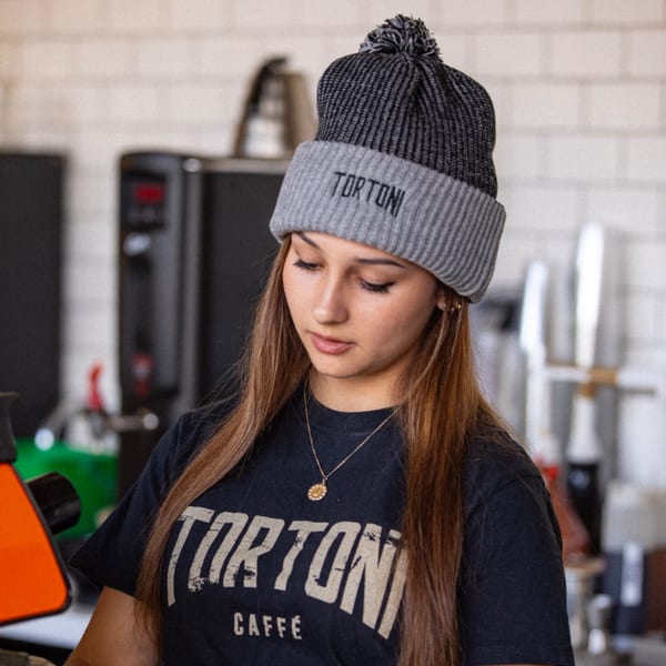 A young woman wearing a Tortoni Caffe beanie and t-shirt, looking down.