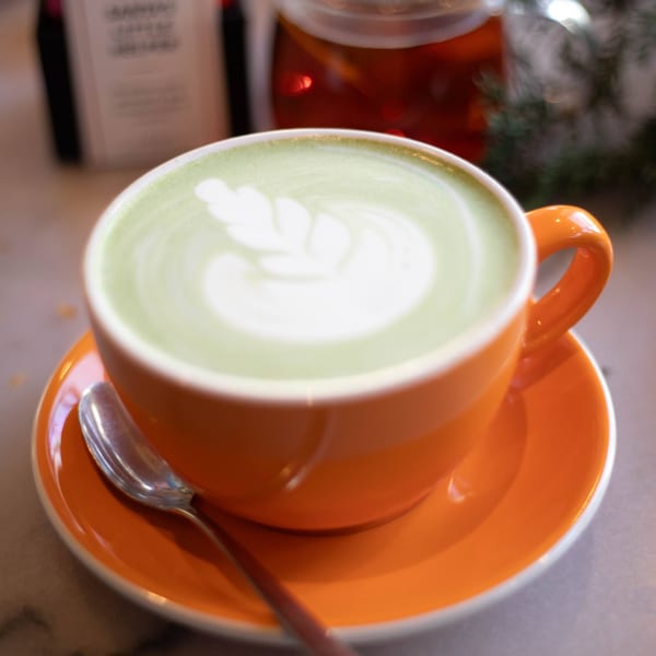 A vibrant orange cup of matcha latte with delicate latte art, a spoon, and a tea pot.