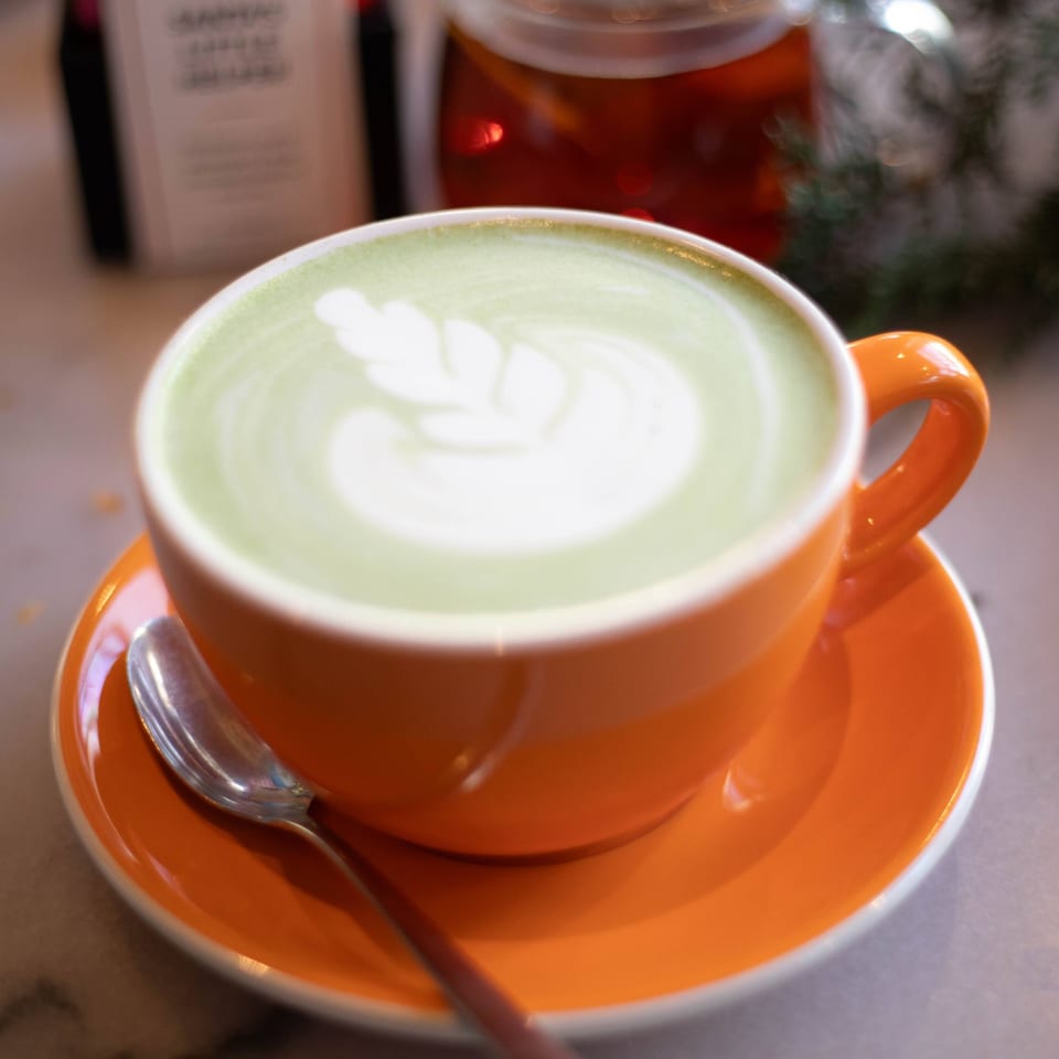 A vibrant orange cup of matcha latte with delicate latte art, a spoon, and a tea pot.