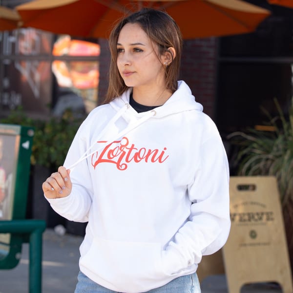 Young woman wearing a white Tortoni Caffe hoodie, standing outdoors with an orange umbrella in background.