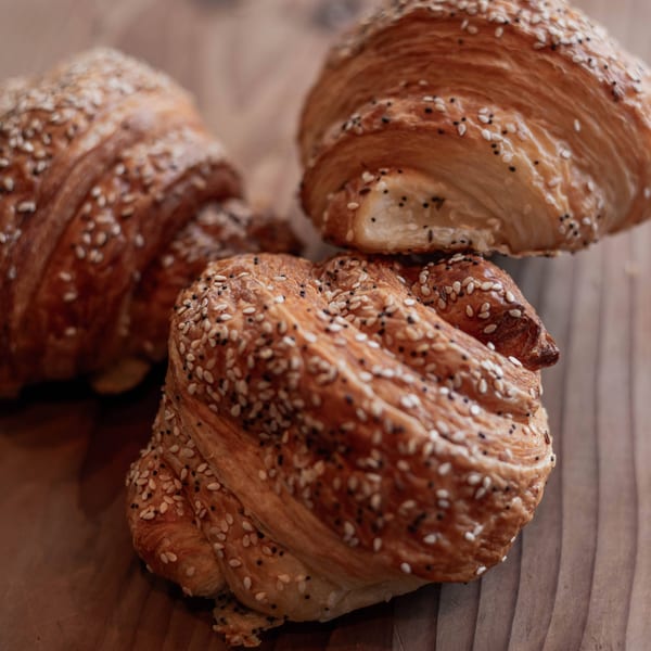 Three golden-brown croissants, generously sprinkled with white sesame and black poppy seeds, on a wooden surface.