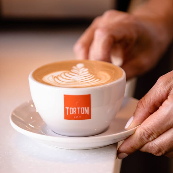 Hands placing a white Tortoni Caffe cup with latte art on a saucer.