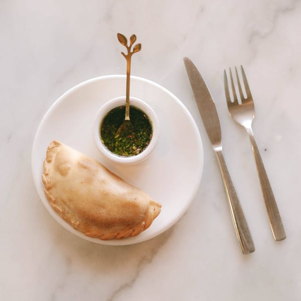 Golden empanada on a white plate with a side of green chimichurri sauce and silverware.