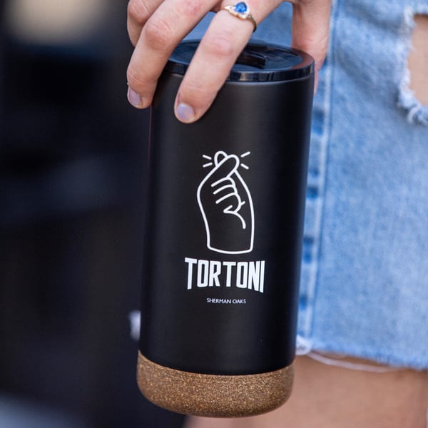 Hand holding a black Tortoni Caffe travel mug with a cork base and white logo.