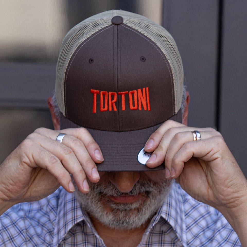 A man wearing a brown Tortoni Caffe hat with red lettering, pulled down over his eyes.
